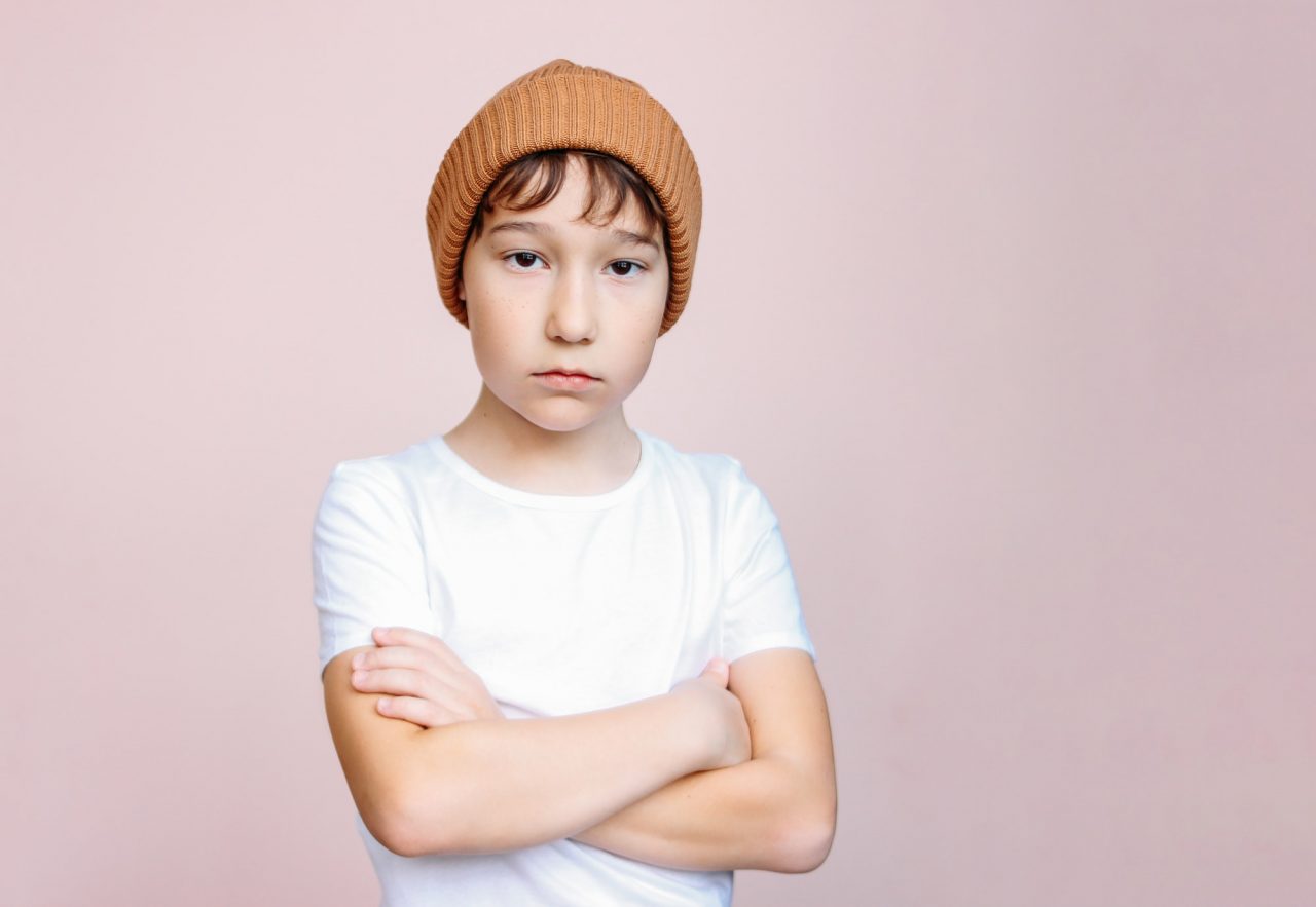 Attractive tween boy with dark hair in hat looks at camera on light pink background.jpg
